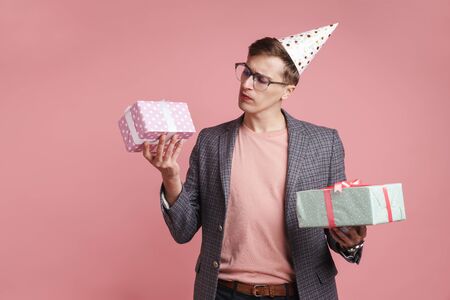 Image of a young thinking serious guy in glasses holding birthday present gift boxes isolated over pink wall background.の写真素材