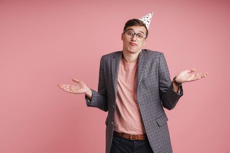 Portrait of an upset young man wearing birthday hat celebrating isolated over pink background, shrugging shouldersの写真素材