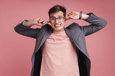 Portrait of an annoyed young attractive man wearing jacket standing isolated over pink background, blocking earsの写真素材