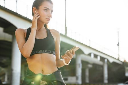 Photo of a concentrated serious young amazing sports fitness woman outdoors listening music with earphones using mobile phone.の写真素材