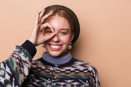Photo of pretty joyful woman in earrings gesturing ok sign and laughing isolated over beige...