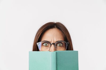 Image of beautiful brunette adult woman wearing eyeglasses frowning and covering her face with book isolated over white backgroundの写真素材