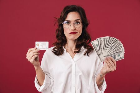 Picture of young confused beautiful woman isolated over red wall background holding money and credit...