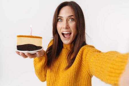 Image of beautiful brunette adult woman wearing sweater holding birthday cake and taking selfie...