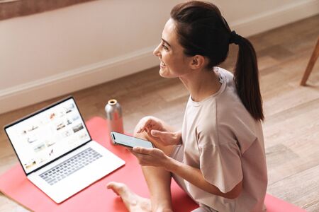 Image of a happy cheery young beautiful brunette fitness woman sit on rug indoors at home near laptop computer using mobile phone.の写真素材