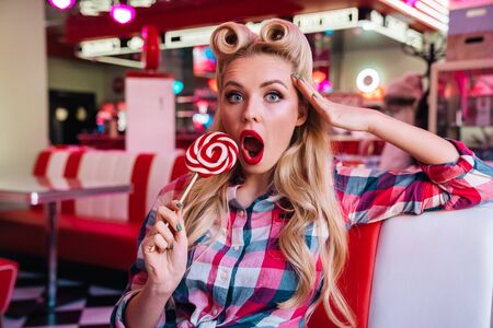 Photo of young seductive woman with beautiful hairstyle holding lollipop and expressing surprise in retro american cafeの写真素材