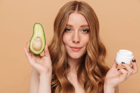 Portrait closeup of gorgeous young half-naked woman with long hair holding avocado and face cream jar isolated over beige backgroundの写真素材