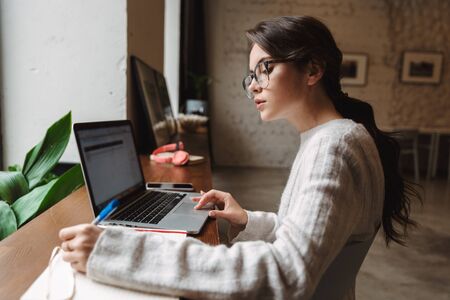 Image of young beautiful caucasian woman wearing eyeglasses using laptop while studying in cafe