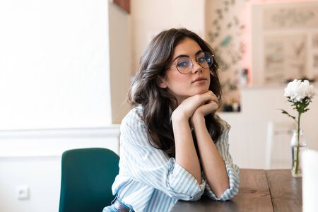 Lovely young brunette woman sitting at the cafe table indoors, relaxing, looking at camera