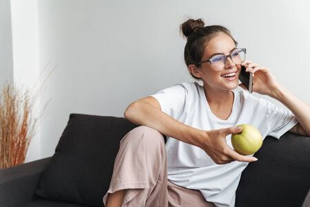 Image of smiling nice woman in eyeglasses talking on cellphone and holding apple while sitting on sofa at living roomの写真素材