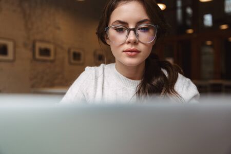 Image of young beautiful caucasian woman wearing eyeglasses using silver laptop in cafeの写真素材
