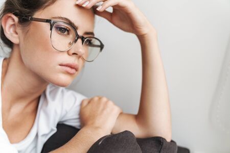 Image closeup of serious cute woman in eyeglasses looking at window while sitting on sofa at living...