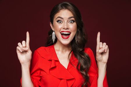 Image of delighted woman wearing red dress pointing fingers at upward isolated over burgundy backgroundの写真素材