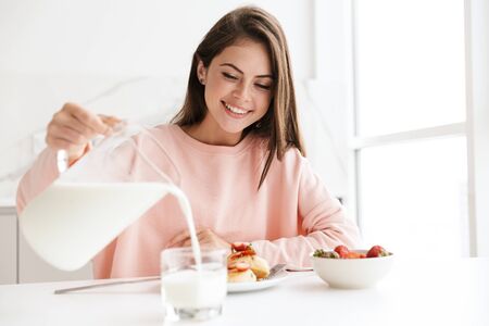 Beautiful smiling young girl having tasty healthy breakfast while sitting at the kitchen tableの写真素材
