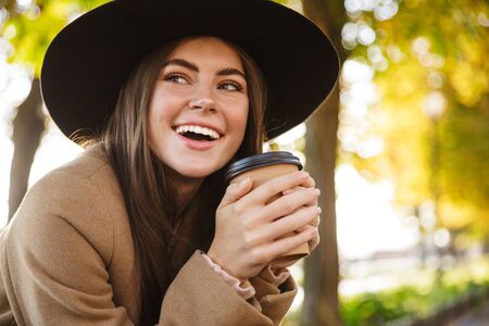 Portrait of happy woman wearing coat and hat drinking coffee while walking in autumn parkの写真素材
