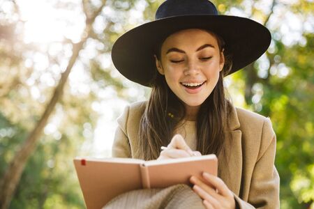 Image of pleased woman in autumn coat and hat holding diary book while sitting on bench in parkの写真素材