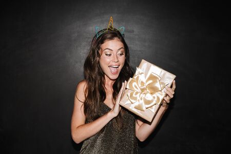 Beautiful happy young girl celebrating isolated over black background, holding present box