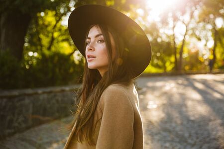Image of brunette woman in autumn coat and hat smiling while walking in green park outdoorsの写真素材
