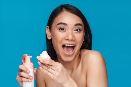 Photo of asian emotional young woman posing isolated over blue wall background holding mousse cleansing foam.の写真素材