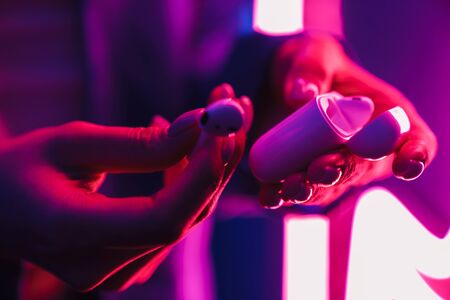 Cropped image of young woman posing over neon lights holding wireless earphones with case.