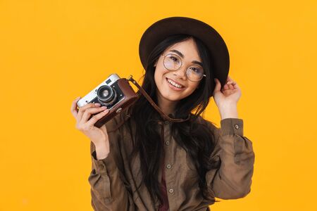 Image of young brunette asian tourist woman wearing hat using retro camera isolated over yellow...