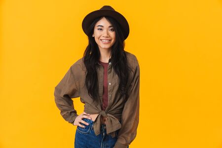 Image of young brunette asian woman wearing hat smiling and looking at camera isolated over yellow backgroundの写真素材