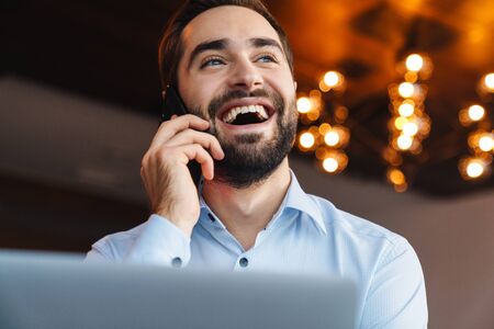 Portrait of handsome young businessman wearing white shirt talking on cellphone and using laptop...