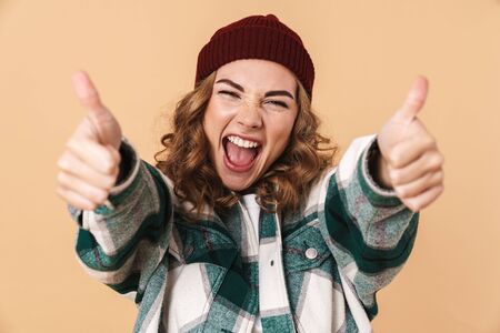 Photo of pretty joyful woman in knit hat laughing and gesturing thumbs up isolated over beige backgroundの写真素材