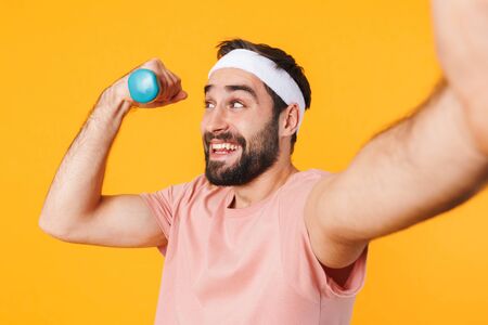 Image of muscular athletic young man in t-shirt taking selfie photo while lifting dumbbell isolated...