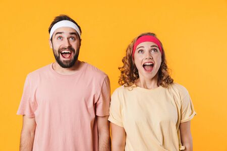 Portrait of athletic young surprised couple wearing headbands expressing excitement and looking upward isolated over yellow backgroundの写真素材