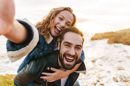 Image of beautiful young caucasian couple smiling and piggybacking while walking by seaside