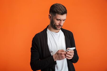 Image of young bearded man wearing basic clothes holding cellphone isolated over orange backgroundの写真素材