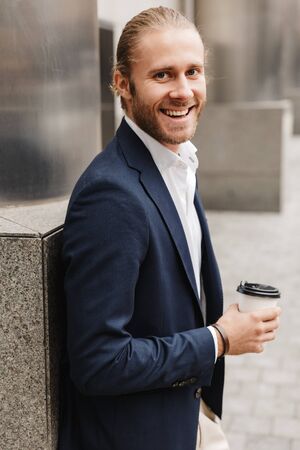 Attractive smiling young blonde haired man in formal wear holding takeaway coffee cup while standing outdoors on a city streetの写真素材