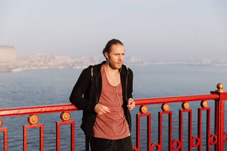 Handsome young fit sportsman listening to music with wireless earphones on a bridge, leaning on railingの写真素材