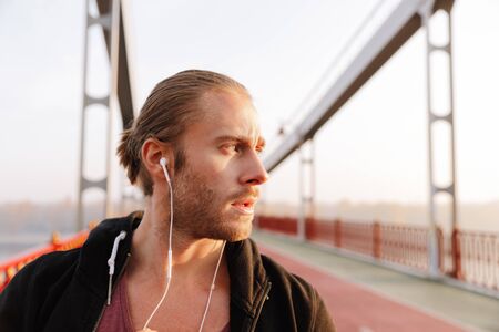 Handsome young fit sportsman listening to music with wireless earphones on a bridge