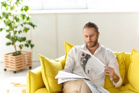 Attractive young blonde haired man wearing white shirt sitting on a couch indoors, reading newspaperの写真素材