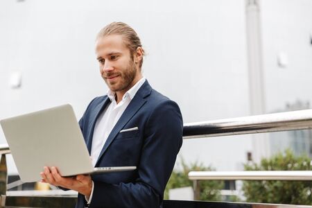 Handsome smiling young bearded businessman standing outdoors in the street, working on laptop computerの写真素材