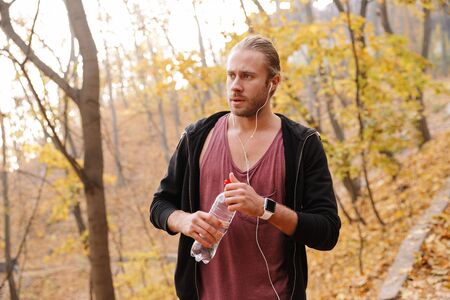 Attractive young fit sportsman jogging in the autumn forest, listening to music with earphones,...