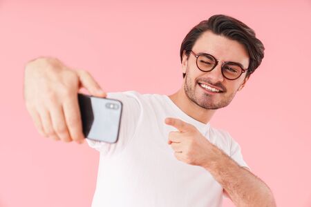 Image of joyful man wearing eyeglasses pointing finger at camera and taking selfie photo on cellphone isolated over pink backgroundの写真素材