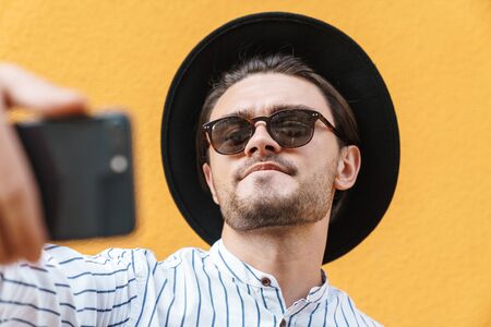Image of young masculine man wearing sunglasses and black hat smiling and taking selfie on cellphone isolated over yellow backgroundの写真素材