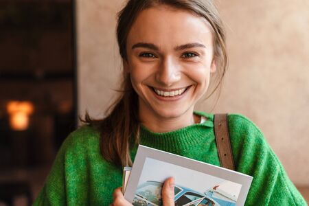 Photo of happy beautiful woman in green sweater holding exercise books and laughing in cozy cafeの写真素材