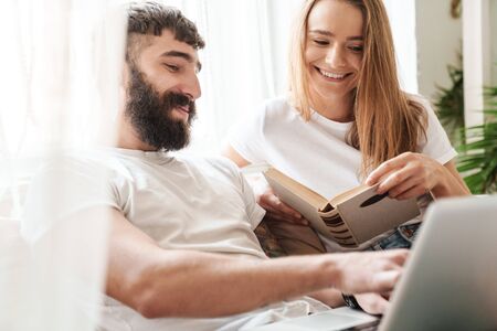 Image of caucasian smiling couple using laptop and reading book while sitting in bright room at home