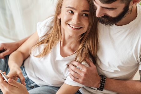 Image of happy young couple smiling and using cellphone while hugging in bright room at homeの写真素材