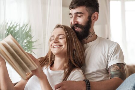 Image of happy young couple laughing and reading book while sitting in bright room at homeの写真素材
