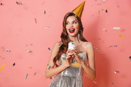 Picture of a young happy woman in bright sequins dress isolated over pink wall background holding cupcake wearing holiday birthday hat over confetti.の写真素材
