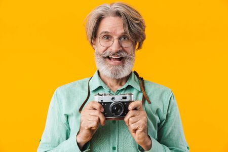 Photo of happy smiling grey-haired man photographer posing isolated over yellow wall background holding camera.の写真素材