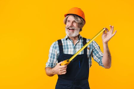 Image of happy emotional elderly grey-haired bearded man builder in helmet posing isolated over yellow wall background holding centimeter.の写真素材