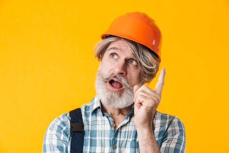 Photo of elderly grey-haired bearded man builder in helmet posing isolated over yellow wall background have an idea.の写真素材