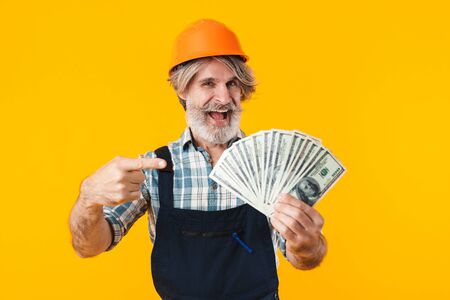 Picture of emotional positive happy elderly grey-haired bearded man builder in helmet posing isolated over yellow wall background holding money pointing.の写真素材