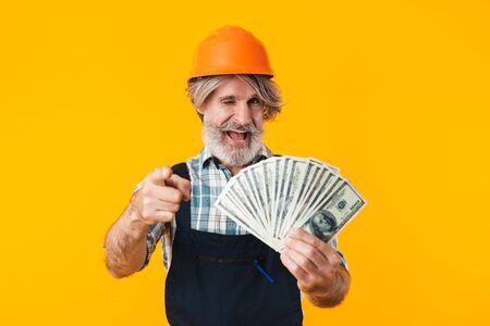 Picture of emotional positive happy elderly grey-haired bearded man builder in helmet posing isolated over yellow wall background holding money pointing to you.の写真素材